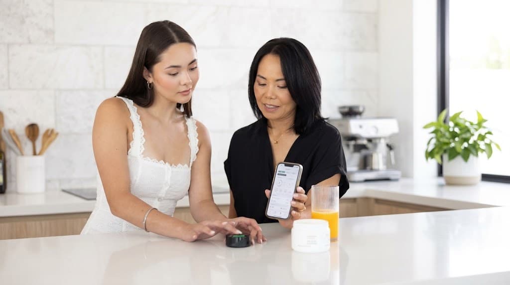 Two women at a kitchen counter, one touching the Prysm iO scanner while the other holds up a phone showing the result